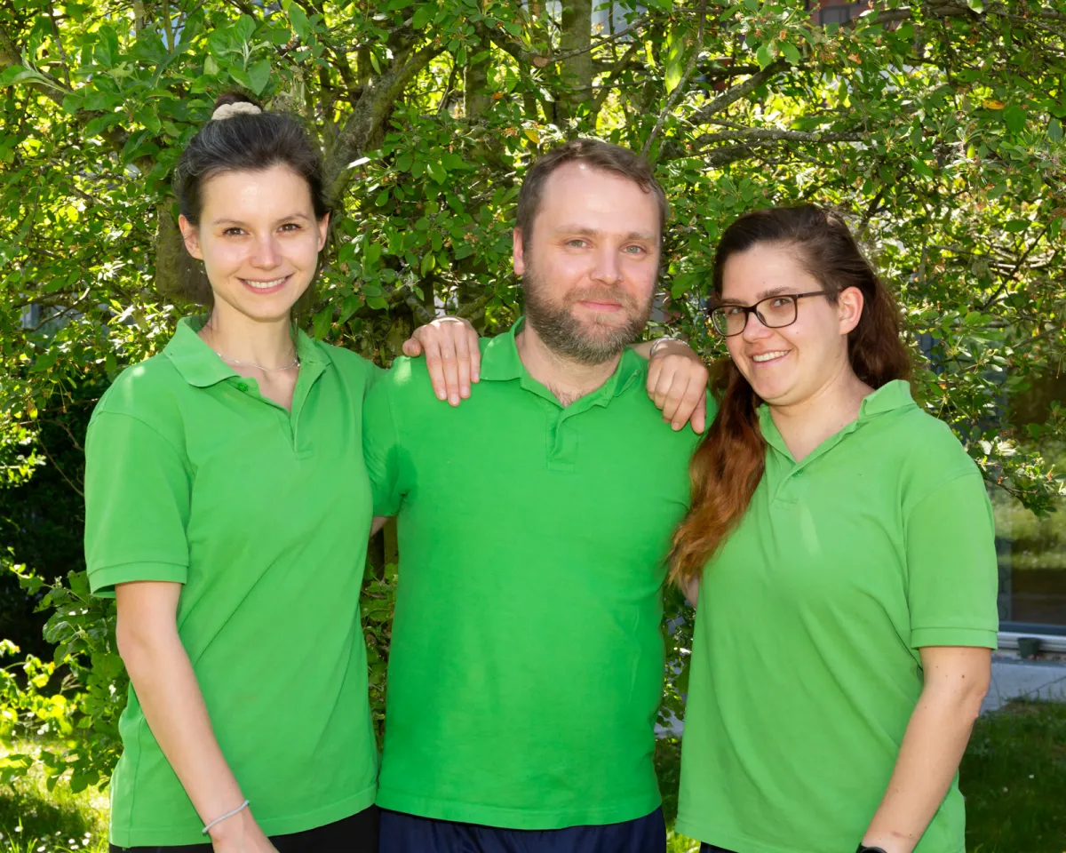 Drei Therapeutinnen und Therapeuten in grünen Poloshirts stehen Arm in Arm vor einem Baum