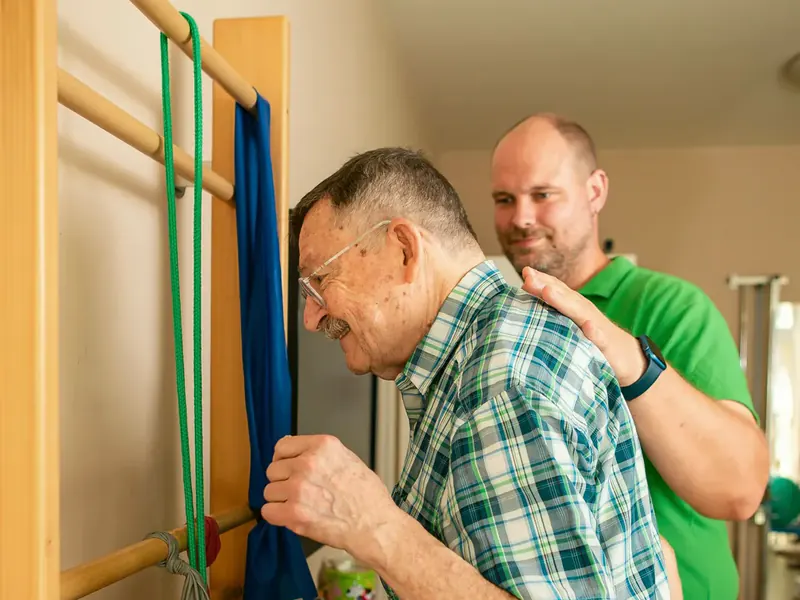 Therapeut begleitet einen älteren Patienten an der Sprossenwand
